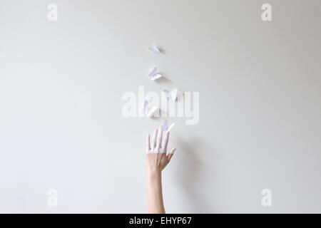 Eine Hand, die Hälfte in weißen Griff nach Papier Schmetterlinge gegen die weiße Wand gemalt Stockfoto