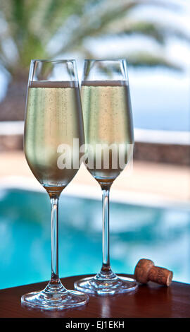 CHAMPAGNERURLAUB zwei frisch gegossene Gläser Champagner und Kork auf einem Terrassentisch im Freien mit luxuriösem Infinity-Pool und Palmen im Hintergrund Stockfoto