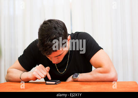 Junger Mann sitzt an einem Tisch in seinem Haus eine Textmitteilung auf seinem Handy mit seinen Fingern zu schreiben, da er mit Freunden kommuniziert Stockfoto