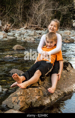 Neunjähriges Mädchen gab ihr sieben Jahre alten Bruder eine Umarmung beim Sitzen auf einem Felsen in einem seichten Fluss Stockfoto