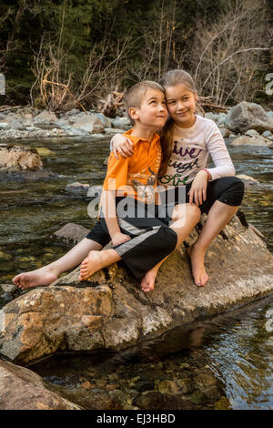 Neunjähriges Mädchen gab ihr sieben Jahre alten Bruder eine Umarmung beim Sitzen auf einem Felsen in einem seichten Fluss Stockfoto