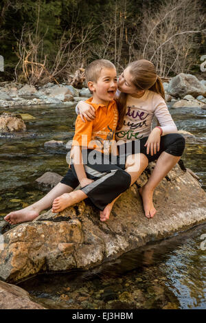 Neunjähriges Mädchen geben ihr sieben Jahre alten Bruder, eine Umarmung und einen Kuss beim Sitzen auf einem Felsen in einem seichten Fluss Stockfoto