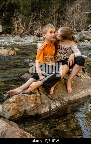 Neunjähriges Mädchen geben ihr sieben Jahre alten Bruder, eine Umarmung und einen Kuss beim Sitzen auf einem Felsen in einem seichten Fluss Stockfoto