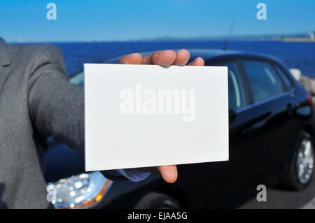 Closeup von einem kaukasischen Jüngling in einem grauen Anzug zeigt ein leeres Schild mit einem schwarzen Auto im Hintergrund Stockfoto
