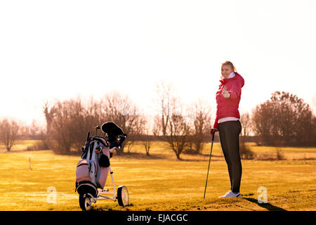 Junge weibliche Golfer in Jacke und Jeans ein Club, während ein Daumen mit Golf-Cart auf der Seite aufgeben. Stockfoto