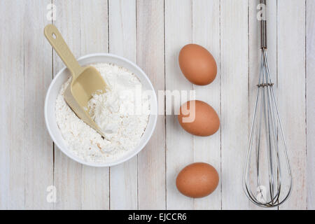 Obenliegende Bild einer Schüssel Mehl, drei braune Eier und einem Metall Schneebesen auf einer rustikalen Holz Küchentisch. Stockfoto