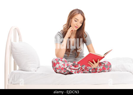 Entspannte Frau im Schlafanzug sitzt auf einem Bett und liest ein Buch isoliert auf weißem Hintergrund, Studioaufnahme Stockfoto