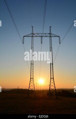 Hochspannungsleitungen mit großen und hohen Pylonen während des Sonnenuntergangs. Die Sonne geht zwischen zwei Pylonen. Stockfoto