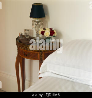 Schwarze Schatten auf Glaslampe mit Vase mit Rosen auf antiken Tisch neben Bett mit weißen Kissen und Bettwäsche Stockfoto