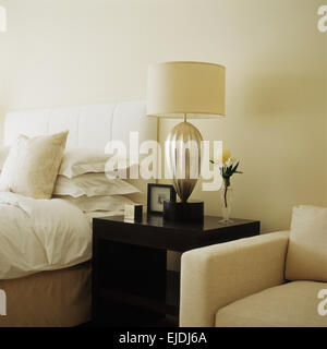 Cremefarbene Sessel neben Schwarz Nachttisch mit Creme Schatten auf Glaslampe in modernen Schlafzimmer mit weißem Leinen auf Polsterbett Stockfoto