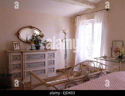 Ovale Spiegel über bemalte Kommode in französischer Landhaus Schlafzimmer mit weißen Voile Vorhänge am Fenster Stockfoto