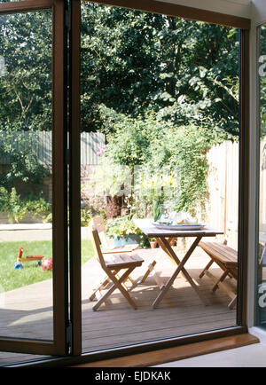 Blick durch offenes Bi-Fold Glas Terrassentüren von Holzmöbeln auf Holzterrasse im Stadtgarten im Sommer Stockfoto