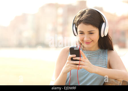 Mädchen Musik hören mit Kopfhörern über ein Smartphone sitzen in einer Bank in einem park Stockfoto