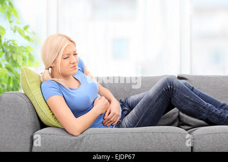 Blonde Frau mit schmerzlichem Gesicht sitzt auf einem grauen Sofa zu Hause mit ihren Händen auf den Bauch gelegt Stockfoto