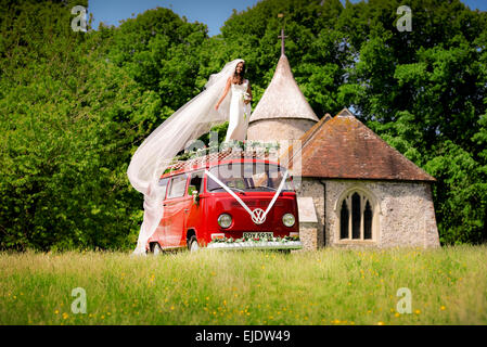 Eine Braut posiert auf dem Dach des hellen roten VW Camper mit Kirche hinter. Stockfoto