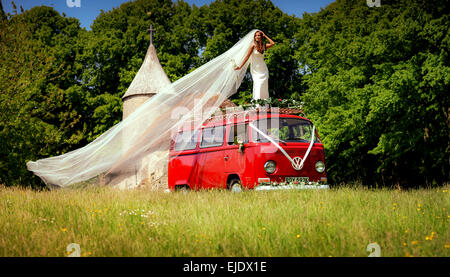 Eine Braut posiert auf dem Dach des hellen roten VW Camper mit Kirche hinter. Stockfoto