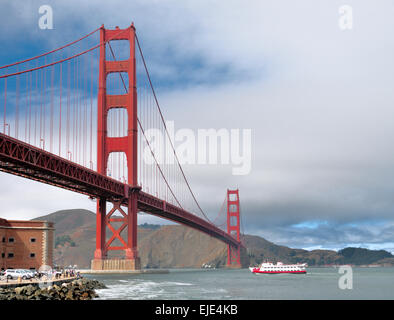 Die legendäre Golden Gate Bridge steht als massive rote Hängestruktur vor einem Hintergrund aus sanften Hügeln und einem bewölkten Himmel, an dem eine Fähre vorbeifährt Stockfoto