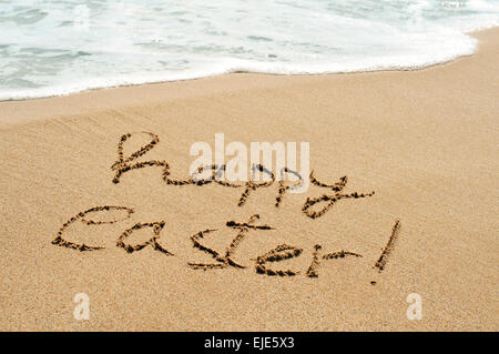 der Satz Frohe Ostern im nassen Sand an der Meeresküste von einem Strand mit einem schäumenden Welle im Hintergrund geschrieben Stockfoto