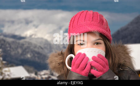 Nettes Mädchen mit einer Tasse Tee im verschneiten Bergdorf, Rauch, Dampf aus heißem Tee mit Berggipfel in der Ferne Stockfoto