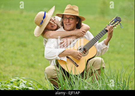 Älteres Paar gemeinsam Spaß haben. Der Mann sitzt und eine Gitarre zu spielen. Die Frau umarmt ihn von hinten. Sie sind im Freien. Stockfoto