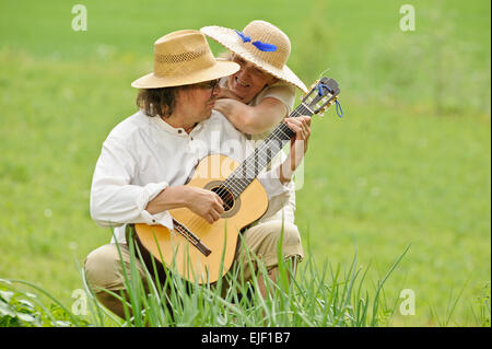 Älteres Paar gemeinsam Spaß haben. Der Mann sitzt und eine Gitarre zu spielen. Die Frau umarmt ihn von hinten. Sie sind im Freien. Stockfoto