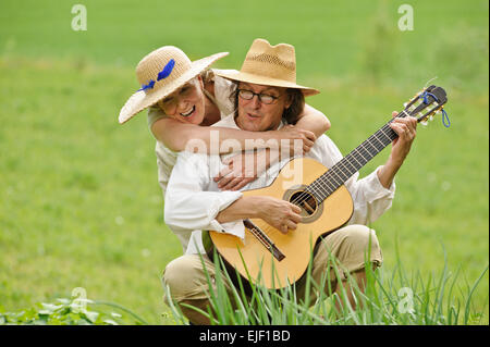 Älteres Paar gemeinsam Spaß haben. Der Mann sitzt und eine Gitarre zu spielen. Die Frau umarmt ihn von hinten. Sie sind im Freien. Stockfoto