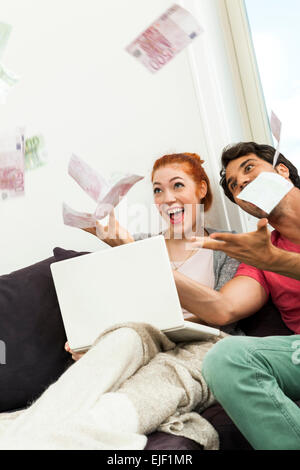 Nahaufnahme sehr glückliches junges Paar mit Laptop sitzen auf der Couch und Euro-Geld in die Luft werfen. Stockfoto