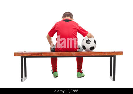 Hintere Ansicht Studio Schuss eines traurigen kleinen Jungen im roten Trikot sitzen auf einer Bank und hält einen Ball isoliert auf weißem Hintergrund Stockfoto