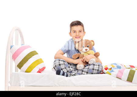 Studioaufnahme von ein fröhlicher kleiner Junge im Pyjama, sitzen im Bett und umarmt einen Teddybär isoliert auf weißem Hintergrund Stockfoto
