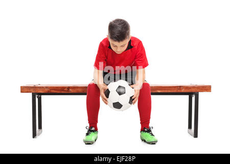 Sorgen kleine Junge im roten Fußballtrikot sitzen auf einer Bank und hält einen Ball isoliert auf weißem Hintergrund Stockfoto