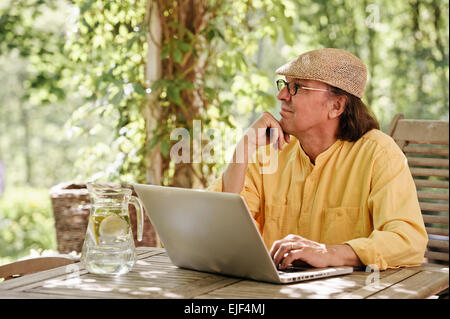 Ältere Mann sitzt im Freien an einem Holztisch unter einer Pergola und funktioniert auf einem Laptopcomputer. Es ist Sommer und es gibt ein Hintergrund Stockfoto
