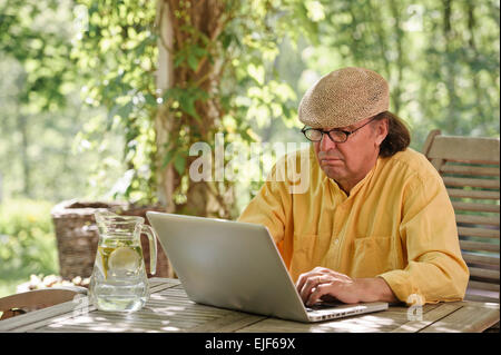 Ältere Mann sitzt im Freien an einem Holztisch unter einer Pergola und funktioniert auf einem Laptopcomputer. Es ist Sommer und es gibt ein Hintergrund Stockfoto