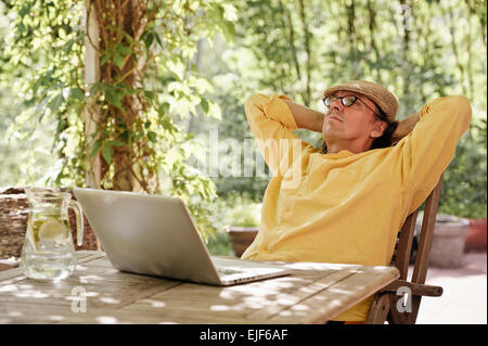 Ältere Mann sitzt im Freien an einem Holztisch unter einer Pergola und funktioniert auf einem Laptopcomputer. Es ist Sommer und es gibt ein Hintergrund Stockfoto
