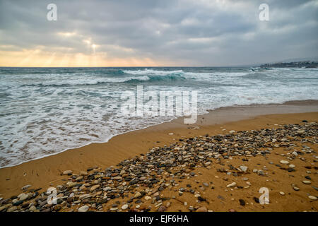 Sonnenuntergang mit Sonnenstrahlen durch die Wolken über stürmische See und leeren Strand Stockfoto