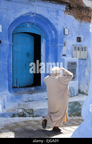 Marokkanischen Mann in traditioneller Kleidung geht in der Nähe von einer blauen Tür in Chefchaouen, Rif-Gebirge, Marokko, Afrika Stockfoto