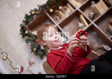 Boy fünf Jahre in Vorfreude auf den Urlaub, in der Nähe der Weihnachtsbaum sitzen hält eine Reihe von roten Beeren Stockfoto