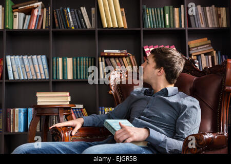 Junger Mann Auschecken Bücher in einer Bibliothek sitzen in einem bequemen Sessel, liest die Titel der Bücher in den Regalen Stockfoto