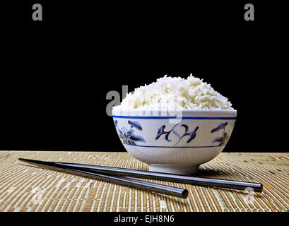 Schüssel mit gekochten Bio Jasmin Reis ungeschält in orientalischen Schüssel auf Gedeck mit Stäbchen auf schwarzem Hintergrund Stockfoto
