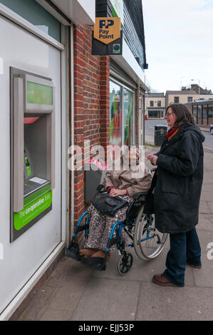 Eine ältere 90 Jahre alte Dame im Rollstuhl ist geholfen, Geld aus einem Geldautomaten Geldautomaten, von ihrem Betreuer oder Helfer zurückziehen. Stockfoto