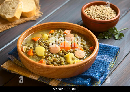 Holzschale Linsensuppe gemacht mit Kartoffel, Karotte, Zwiebel und Wurst Scheiben, mit eine kleine Schüssel mit rohem Linsen in den Rücken Stockfoto