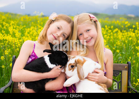 Zwei schöne Schwestern halten Hasen sitzend auf einer Bank in einem Feld von gelben Blüten. Stockfoto