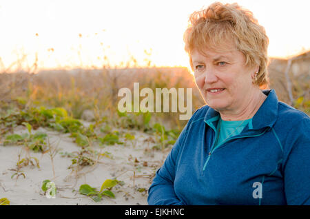 Senioren weiblich-Alter 60-65 stehend mit der Sonne auf dem Rücken in den Dünen in Strandnähe suchen, entspannt und glücklich Stockfoto