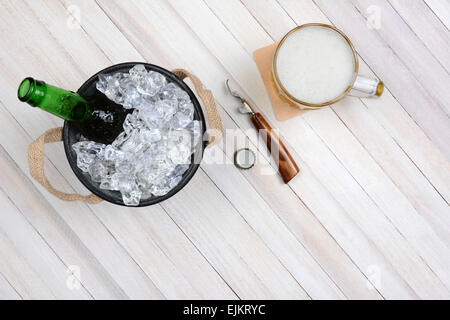 Overhead Schuss von einem Eiskübel mit einer geöffneten Bierflasche einen Becher Bier und Opener auf einem rustikalen weißer Holztisch. Horizontal für Stockfoto