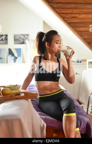 Gesunden Lebensstil: eine slim-fit attraktive junge asiatische ethnische chinesische Mädchen Trägerin Sportbekleidung entspannen, trinken ein Glas kalte Milch nach dem Training in ihrem Schlafzimmer zu Hause Stockfoto