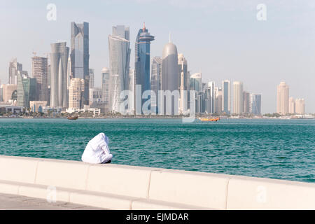 Eine Qatari Mann sitzt allein auf der Stadt am Wasser mit den Wolkenkratzern von Doha im Hintergrund. Stockfoto