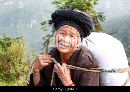 Lächelnde ältere Frau der Miao-Minderheit schweren Sack Reis auf dem Rücken, an der Bergstraße in der ländlichen Provinz Guizhou Stockfoto