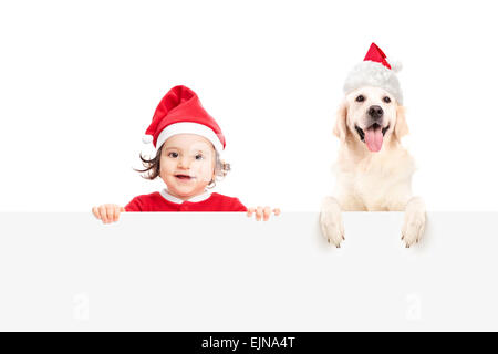 Babymädchen im Weihnachtsmann-Kostüm und ein Hund mit Weihnachtsmann Mütze posiert hinter einer leeren weißen Plakatwand Stockfoto