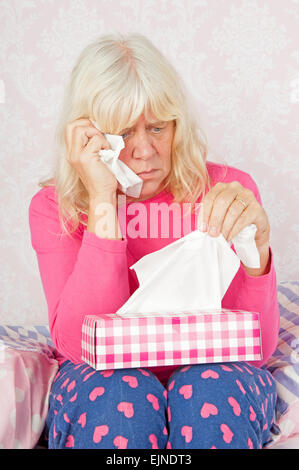 Traurige Frau mit rosa Pyjama und Gewebe auf Boden gegen Bett sitzend Stockfoto