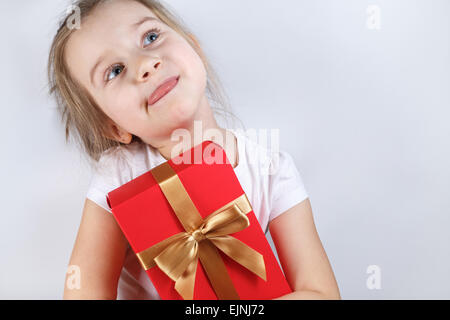 Kleines Mädchen mit einer roten Geschenkbox mit goldenen Band gebunden Bogen. Kind streckte seine Zunge mit Vergnügen. Platz für Text. Stockfoto