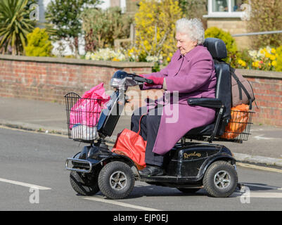 Mobility Scooter. Ältere Frau reiten ein Mobility Scooter über eine Straße, die einkaufstaschen in England, Großbritannien. Stockfoto